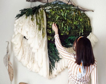 May include: A large, white, fluffy, yarn wall hanging with a green and brown woven tapestry section. The tapestry features a landscape design with a branch and leaves. A person is adjusting the tapestry.