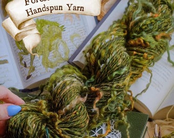 Pode incluir: Um novelo de fio fiado à mão em tons de verde, castanho e laranja, com o texto "Forest Roots Handspun Yarn" numa faixa. O fio é exibido em um livro aberto com ilustrações e um pedaço de madeira coberto de musgo.