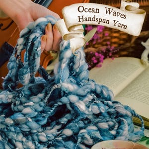 May include: A close-up of Ocean Waves Handspun Yarn in shades of blue and white. The thick yarn is held in a hand, with a violin and open book in the background. A teacup with a floral design is in the foreground.