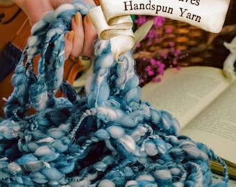 Pode incluir: Close-up de Ocean Waves Handspun Yarn em tons de azul e branco. O fio grosso é segurado em uma mão, com um violino e um livro aberto ao fundo. Uma xícara de chá com um design floral está em primeiro plano.