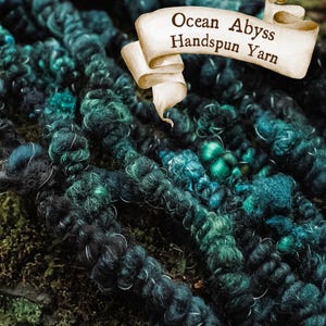 Può includere: Primo piano di filato filato a mano nei toni del verde acqua, verde e nero. Il filato è spesso e strutturato, con un aspetto leggermente sfocato. Un banner recita "Ocean Abyss Handspun Yarn".