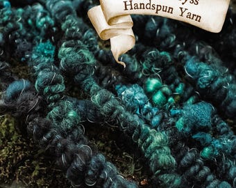 Pode incluir: Close-up de fio fiado à mão em tons de verde-azulado, verde e preto. O fio é espesso e texturizado, com uma aparência ligeiramente difusa. Uma faixa diz "Ocean Abyss Handspun Yarn".