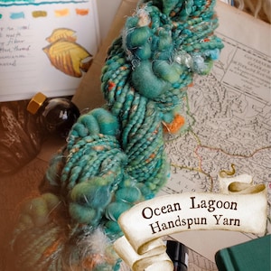 May include: A close-up of a skein of Ocean Lagoon handspun yarn. The yarn is a mix of teal, turquoise, and orange. The image also includes a vintage map, ink bottle, and a sketch of a tree.