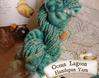 Pode incluir: Uma aproximação de um novelo de fio fiado à mão Ocean Lagoon. O fio é uma mistura de verde-azulado, turquesa e laranja. A imagem também inclui um mapa vintage, um frasco de tinta e um esboço de uma árvore.