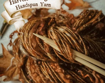 Pode incluir: Um close-up de um novelo de fio fiado à mão em tons de castanho e dourado, com o texto "Harvest Foliage Handspun Yarn" numa faixa. Duas agulhas de tricô de madeira estão colocadas sobre o fio, com folhas secas no fundo.