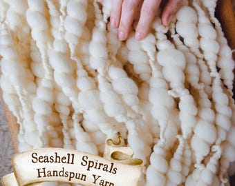 May include: Cream-colored, handspun yarn with a seashell spiral design. The thick yarn is arranged in a textured pattern. The image includes a banner that reads "Seashell Spirals Handspun Yarn".