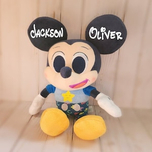 Pu&ograve; includere: Un giocattolo di peluche di Topolino con orecchie nere e una camicia blu. Le orecchie sono ricamate con i nomi "Jackson" e "Oliver".