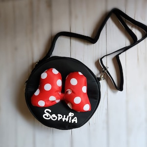 May include: Black round crossbody bag with a red and white polka dot bow and the name "Sophia" written in white on the front.