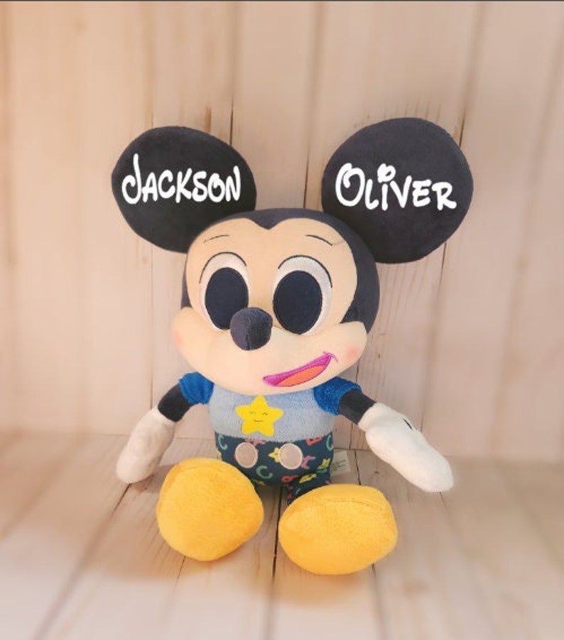 Pu&ograve; includere: Un giocattolo di peluche di Topolino con orecchie nere e una camicia blu. Le orecchie sono ricamate con i nomi "Jackson" e "Oliver".