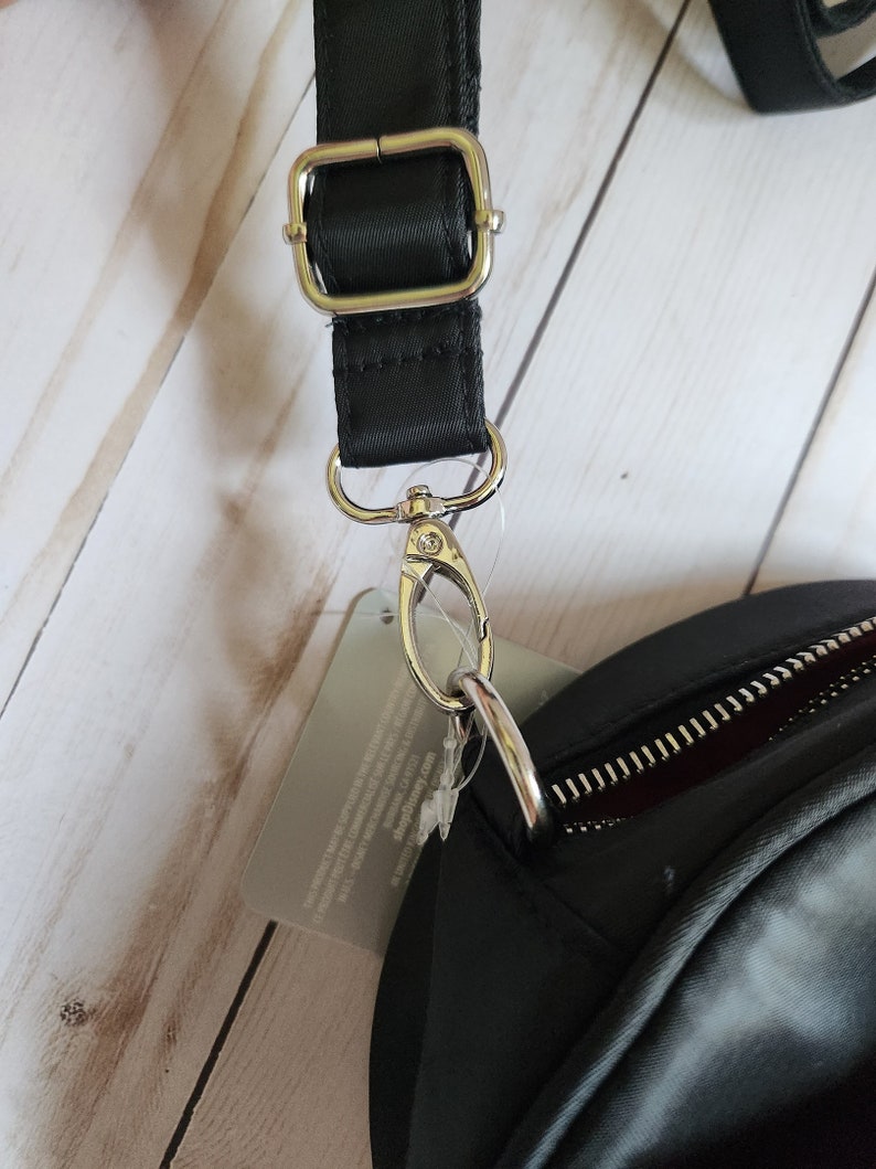 May include: A black crossbody bag with a silver zipper and a silver clasp. The bag has a black adjustable strap with a silver buckle.