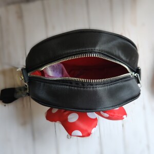 May include: Black crossbody bag with a red and white polka dot bow. The bag has a zipper closure and a silver zipper pull.
