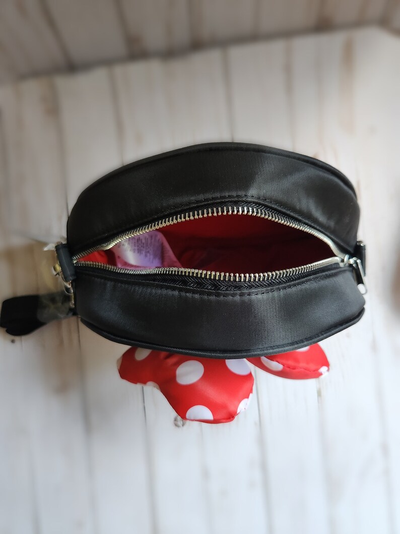 May include: Black crossbody bag with a red and white polka dot bow. The bag has a zipper closure and a silver zipper pull.