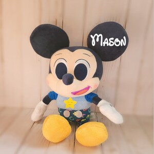 Pu&ograve; includere: Un giocattolo di peluche di Topolino che indossa una camicia blu con una stella gialla e lettere colorate. Il giocattolo ha grandi occhi neri e una bocca sorridente. L'orecchio destro del giocattolo ha il nome "MASON" scritto su di esso.