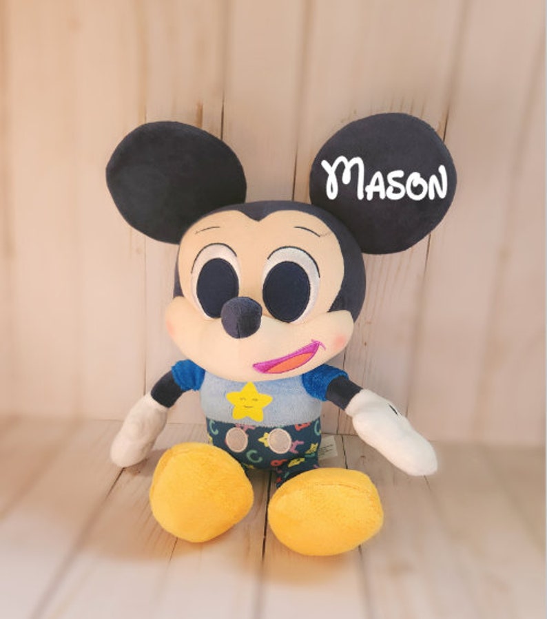 Pu&ograve; includere: Un giocattolo di peluche di Topolino che indossa una camicia blu con una stella gialla e lettere colorate. Il giocattolo ha grandi occhi neri e una bocca sorridente. L'orecchio destro del giocattolo ha il nome "MASON" scritto su di esso.