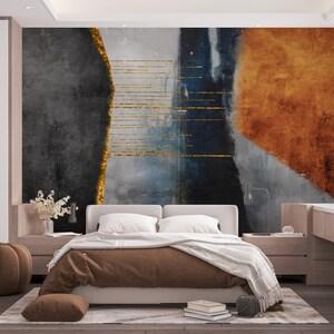May include: A bedroom interior with a large abstract wall mural featuring shades of gray, blue, and orange with gold accents. The mural is behind a white bed with brown bedding and a white throw blanket. There is a brown ottoman in the foreground.