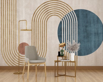 Op de afbeelding: Een woonkamer interieur met een beige muur met een geometrisch patroon van gouden en blauwe cirkels en bogen. Een grijze stoel en een gouden bijzettafel met een vaas met gedroogde bloemen staan op de voorgrond.