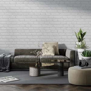 May include: A living room with a green sofa, two coffee tables, a green ottoman, and a white brick wall. The coffee tables are made of glass and metal. The ottoman is covered in fabric. The sofa has a pillow with a floral pattern.