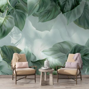 May include: Two beige armchairs with wooden frames are positioned in front of a large, green leaf mural. A small, round table with a decorative vase and bowl sits between the chairs. Pillows and blankets add to the cozy aesthetic.