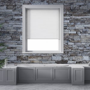 May include: A white bathtub with a gray tiled surround and a window with white blinds. The wall behind the tub is made of stacked stone tiles in shades of gray and brown.