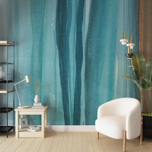 May include: A teal and turquoise watercolor-style wall mural with vertical brushstrokes. The room features a white armchair, a small wooden side table with a lamp and vase, and a black metal bookshelf. The floor is light wood.