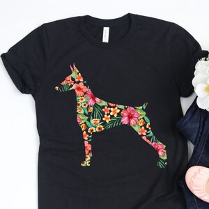 May include: A black t-shirt with a floral print of a Doberman Pinscher. The shirt is laying on a white surface with a pair of blue jeans and pink flats.