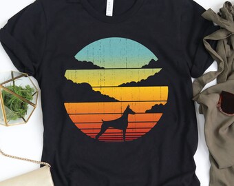 Può includere: T-shirt nero con un design retrò del tramonto e la silhouette di un cane. La grafica presenta un gradiente dal blu all'arancione, con il cane in nero. La maglietta è abbinata a pantaloni verde oliva e occhiali da sole.