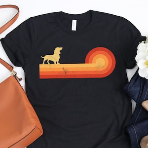 May include: Black t-shirt featuring a dachshund silhouette above a retro sunset design in shades of orange and yellow. The shirt is styled with a brown purse, blue jeans, and beige flats.