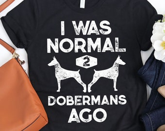 Op de afbeelding: Zwart T-shirt met witte tekst: "I WAS NORMAL 2 DOBERMANS AGO." Het ontwerp toont twee Dobermann-silhouetten. Het shirt wordt getoond met een bruine handtas, een blauwe jeans en beige ballerina's.