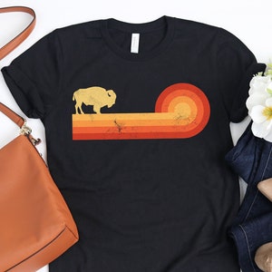 May include: Black t-shirt featuring a retro design with a bison silhouette and a sunset graphic in shades of orange and red. The shirt is paired with blue jeans, tan shoes, and a brown purse.
