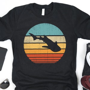 May include: A black t-shirt with a vintage-style graphic of a shark silhouette swimming in front of a sunset. The sunset is depicted with a blue, teal, orange, yellow, and red color scheme.