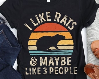 May include: Black t-shirt with the text "I LIKE RATS & MAYBE LIKE 3 PEOPLE" in white. The design includes a rat silhouette against a sunset background with orange, yellow, and teal stripes. The shirt is paired with blue jeans, sandals, and a gray sweater.
