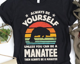 May include: Black t-shirt featuring the text "ALWAYS BE YOURSELF UNLESS YOU CAN BE A MANATEE THEN ALWAYS BE A MANATEE" in white, with a manatee silhouette. The design is set against a sunset-style background. The shirt is styled with olive green trousers and sunglasses.