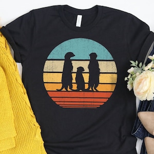 May include: A black t-shirt with a retro sunset graphic featuring three meerkats. The meerkats are silhouetted against a blue, orange, and yellow sunset.