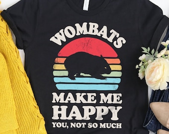 May include: Black t-shirt featuring a wombat silhouette and the text "WOMATS MAKE ME HAPPY YOU, NOT SO MUCH". The design includes a retro sunset graphic. The shirt is styled with a mustard yellow cardigan, denim jeans, and shoes.