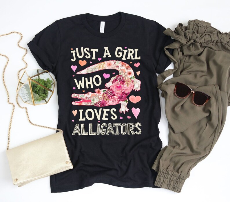 Puede incluir: Camiseta negra con el texto "JUST A GIRL WHO LOVES ALLIGATORS". El gr&aacute;fico del caim&aacute;n est&aacute; lleno de patrones florales rosas y blancos. La imagen tambi&eacute;n incluye pantalones verde oliva, gafas de sol y un clutch dorado.