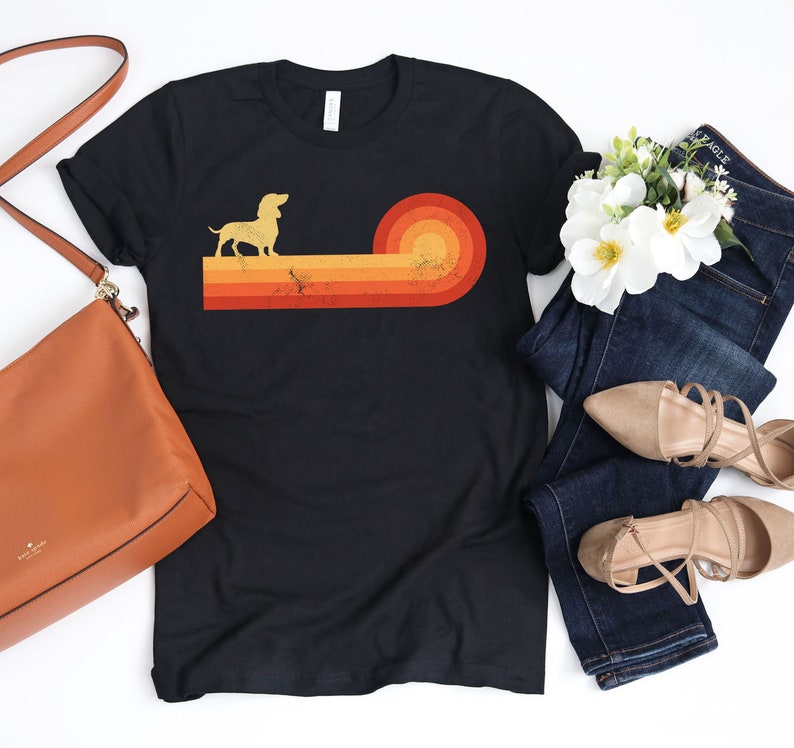 May include: Black t-shirt featuring a dachshund silhouette above a retro sunset design in shades of orange and yellow. The shirt is styled with a brown purse, blue jeans, and beige flats.