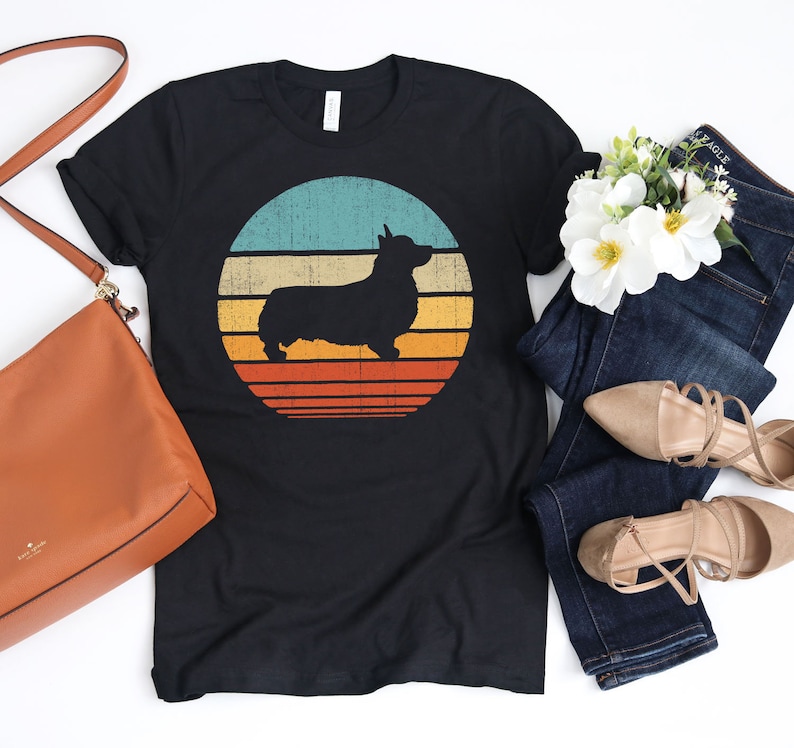 Puede incluir: Una camiseta negra con un gr&aacute;fico de estilo vintage de una silueta de corgi frente a una puesta de sol. El gr&aacute;fico est&aacute; en tonos de azul, amarillo y naranja.