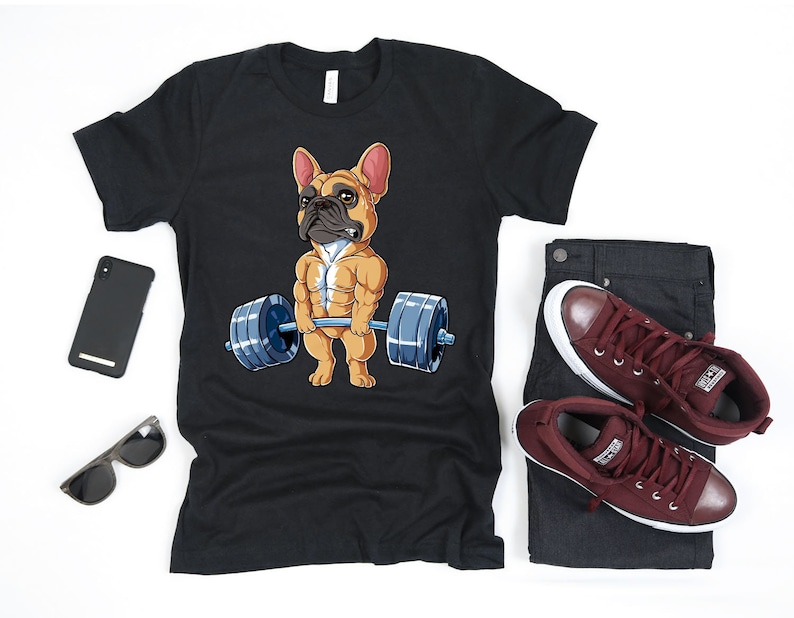 May include: Black t-shirt featuring a cartoon image of a French bulldog lifting weights. The bulldog is wearing a red collar and has a muscular physique.