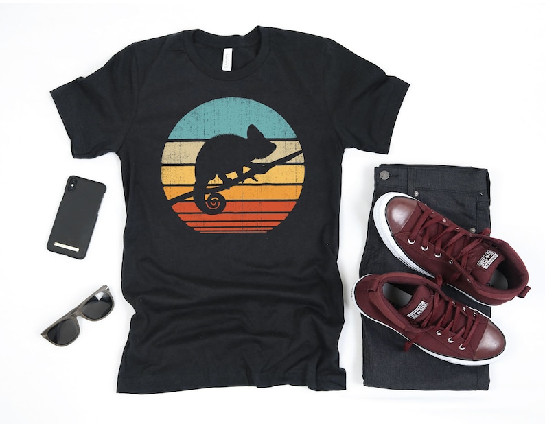 May include: A black t-shirt with a vintage-style graphic of a chameleon on a branch in front of a sunset. The graphic is in shades of blue, orange, yellow, and red.