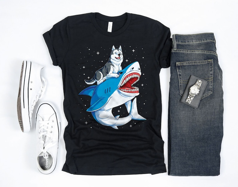 K&ouml;nnte beinhalten: Ein schwarzes T-Shirt mit einem Aufdruck eines Husky-Hundes, der auf einem Hai im Weltraum reitet. Der Hai ist blau und wei&szlig; mit einem gro&szlig;en, offenen Maul. Der Husky ist schwarz und wei&szlig; mit blauen Augen. Der Hintergrund ist schwarz mit wei&szlig;en Sternen.