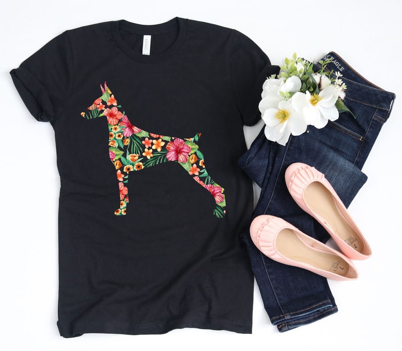 May include: A black t-shirt with a floral print of a Doberman Pinscher. The shirt is laying on a white surface with a pair of blue jeans and pink flats.