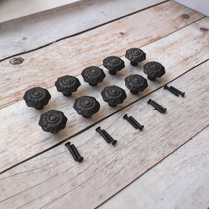 May include: A set of ten round, matte black cabinet knobs featuring a raised, floral-inspired petal design on the surface. Each knob is accompanied by a matching black metal mounting screw, arranged in a grid pattern for furniture hardware.