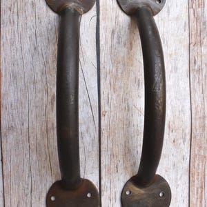 May include: Two antique-style, dark brown metal door handles. Each handle has a circular base with three screw holes and a curved, cylindrical grip. The handles are mounted on a light brown wooden surface.