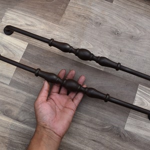 Könnte beinhalten: Zwei dunkelbraune, dekorative Schrankgriffe. Jeder Griff hat ein zentrales, bauchiges Design mit einer Stange, die zu kreisförmigen Enden führt. Die Griffe sind aus Metall und für Möbel bestimmt. Die Griffe sind auf einer Holzmaserungsoberfläche abgebildet.