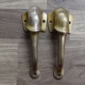 May include: Two antique brass door handles featuring elephant head designs. The handles have a textured, chevron pattern and are mounted on a wooden surface.