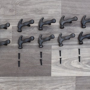 Könnte beinhalten: Zwölf schwarze Metallhaken in Hammerform mit zwölf schwarzen Schrauben. Die Haken sind in drei Reihen zu vier auf einer Holzoberfläche angeordnet.