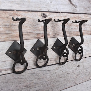 Puede incluir: Cuatro ganchos de pared de metal gris oscuro, cada uno con una placa de montaje en forma de diamante con dos orificios para tornillos, un gancho curvo hacia arriba y un anillo circular unido a la base. Los ganchos están en una fila.