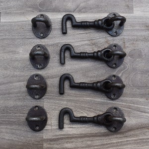May include: Four sets of black iron gate hardware, including hook and eye latches and matching oval plates. The hardware is arranged on a grey wood-grain surface. The hooks are curved and the eyes are circular.