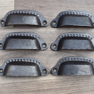 Könnte beinhalten: Sechs schwarze Schubladengriffe aus Gusseisen mit einem dekorativen Perlenrand. Die Griffe sind alle gleich groß und gleich geformt und haben ein leicht geschwungenes Design.