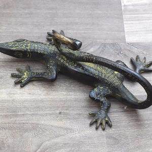 Può includere: Statuetta di lucertola verde scuro e oro dettagliata. Il rettile ha un corpo strutturato con accenti gialli ed è posizionato su una superficie di legno. La coda della lucertola si curva attorno al suo corpo.